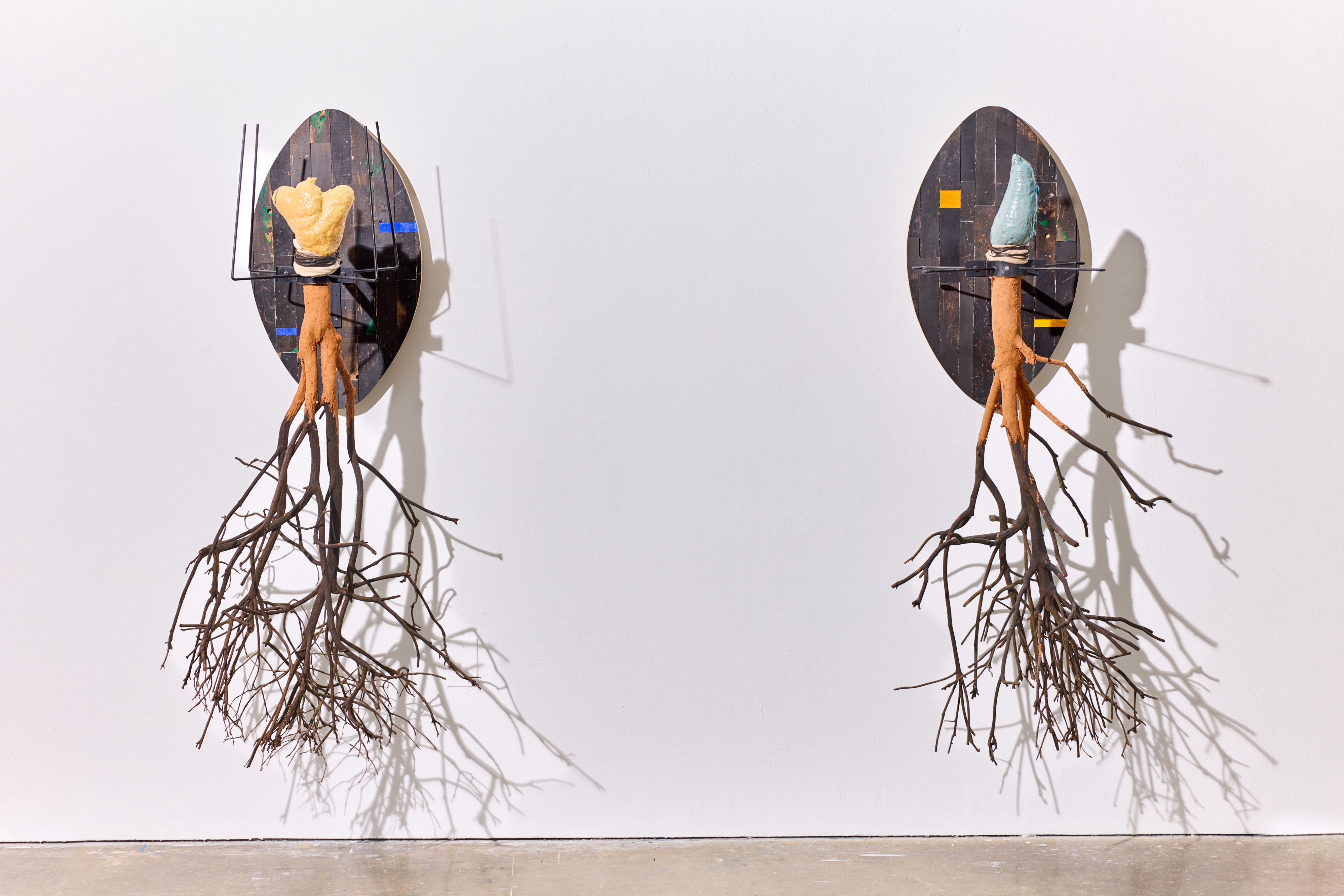 The Oracle and the Conjurer, 2024 — two oval wall-mounted sculptures with figures emerging from charred wood panels and roots descending to the floor